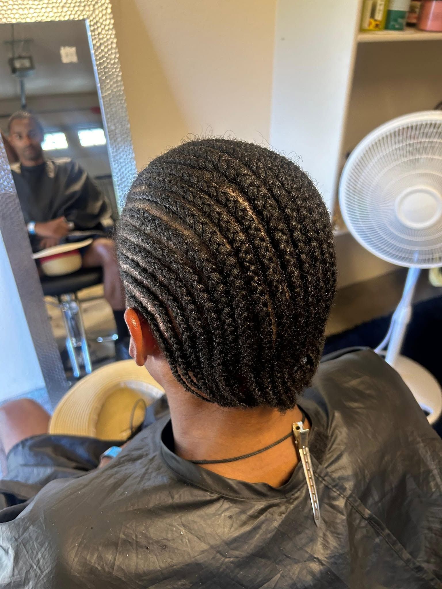Person with short patterned hair sitting in a salon chair, seen from behind beside a standing fan.