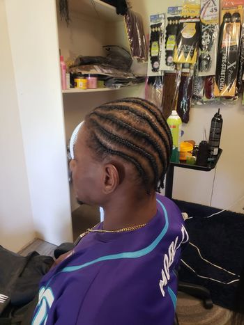 Person with braided hair seated in a salon chair, wearing a purple cape, with hair tools on the wall behind.