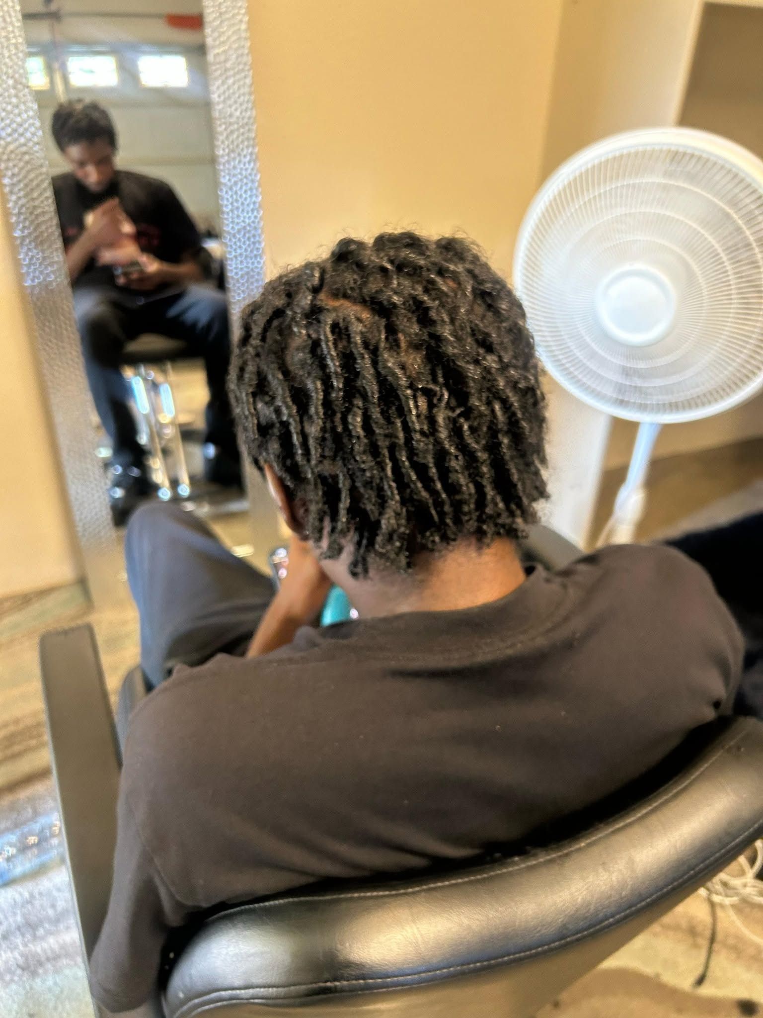 Person seated in a salon chair, seen from behind, with a mirror reflection and a standing fan nearby.