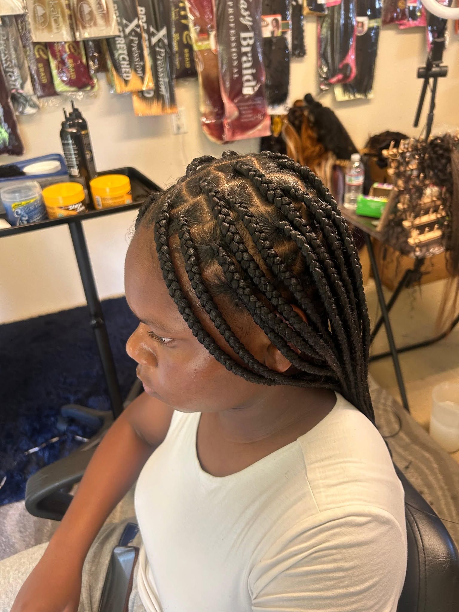 Person with braided hair sits in a salon chair, viewed from the side, with hair products and braids in the background.