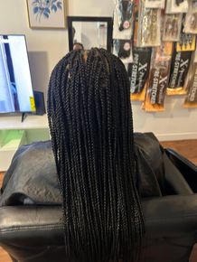 Person with long black braids seated in a salon chair, facing a wall of hair extensions.