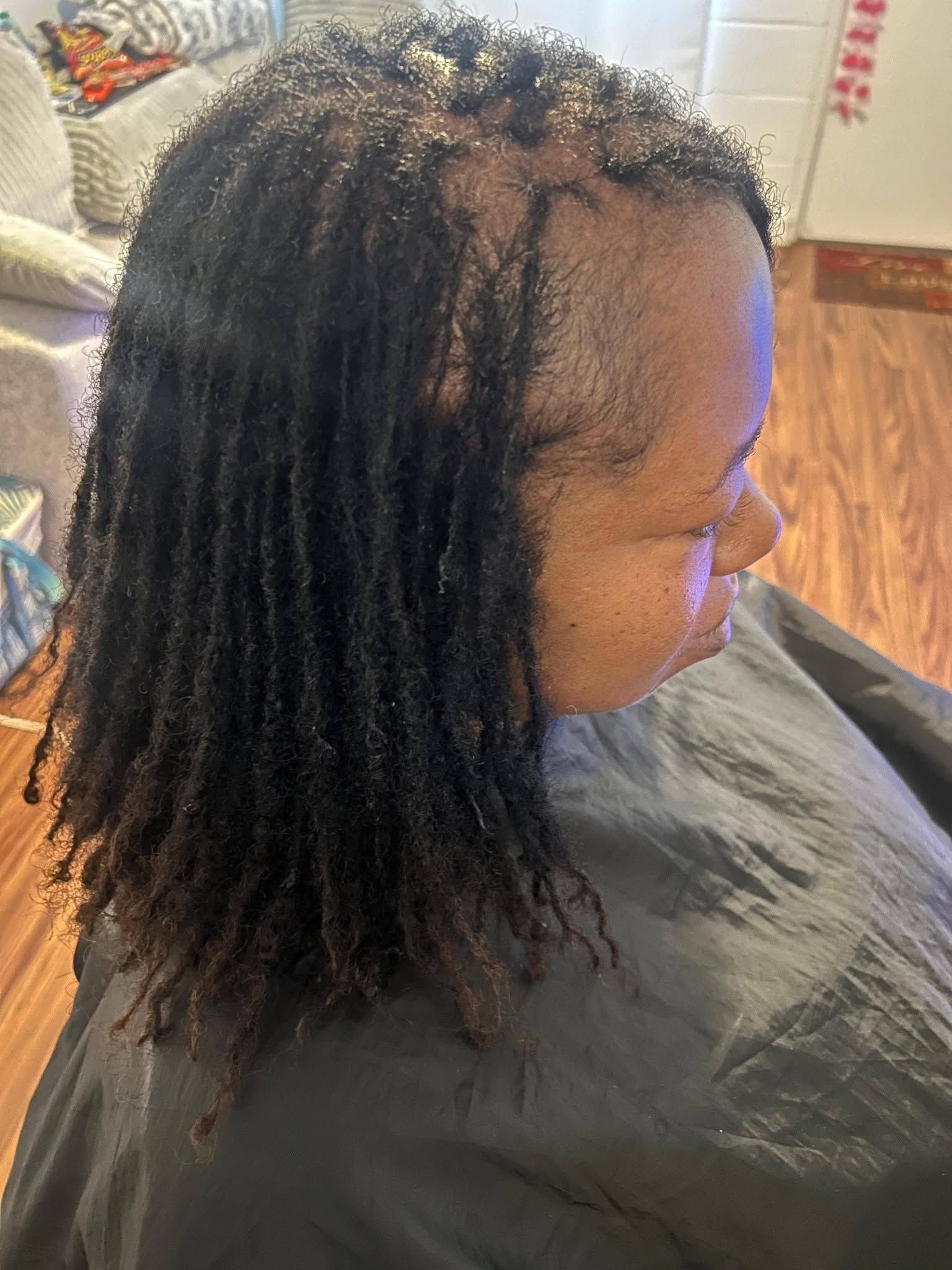 Person with shoulder-length locs seated indoors, shown in profile wearing a dark cape or smock.