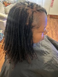 Person with shoulder-length locs seated indoors, shown in profile wearing a dark cape or smock.