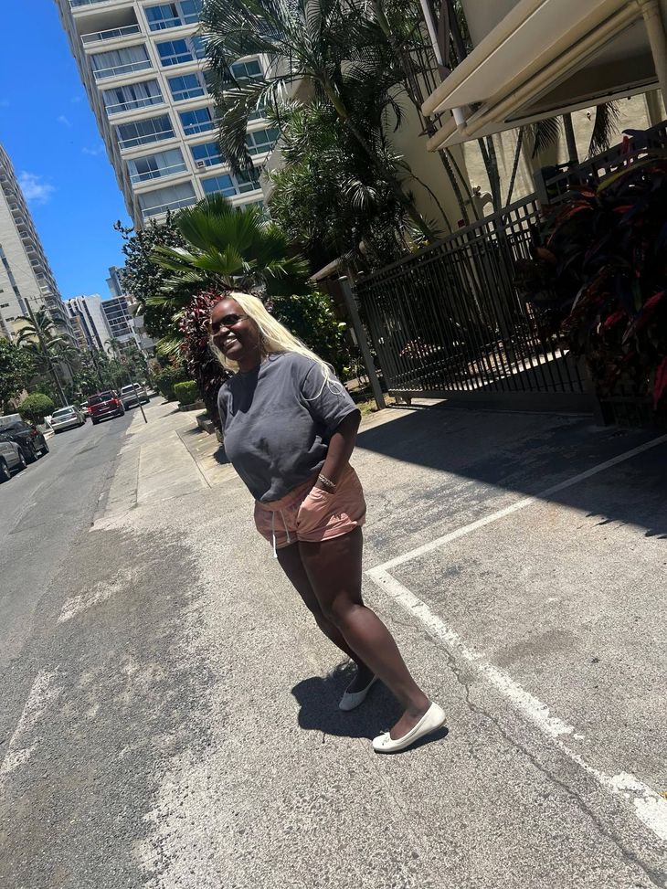Woman standing on a sunny city street beside a tall building and fence, wearing a gray top and pink shorts.