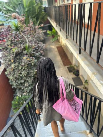Person walking down outdoor stairs beside black railing and lush greenery, carrying a pink bag.
