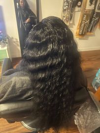 Long wavy black hair seen from behind in a salon chair, with a mirror and wall decor in the background.
