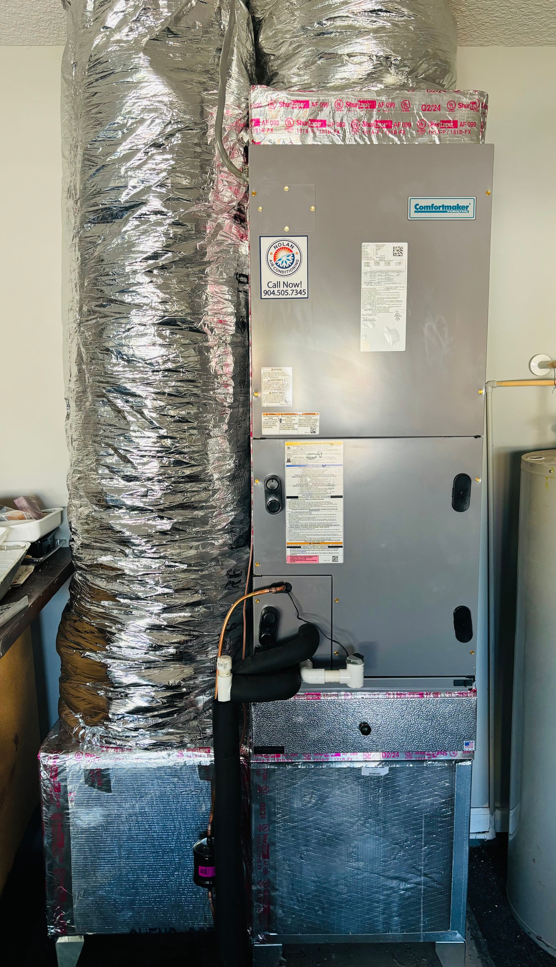 A silver air conditioner is sitting in a room next to a water heater.