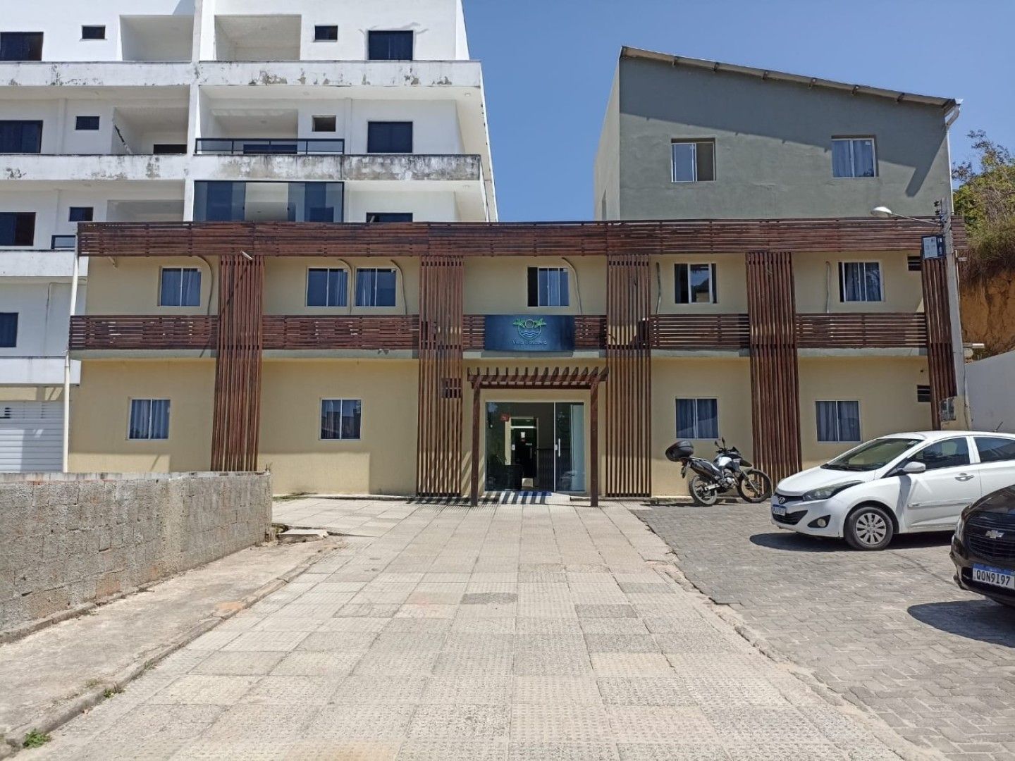 Fachada de edifício moderno com paredes cinzentas, coluna de tijolo e treliça no telhado contra um céu azul.