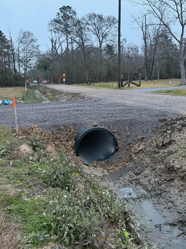 A dirt road with a large culvert pipe under it, surrounded by trees and muddy drainage ditch.