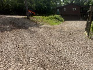 Gravel driveway leading to a brown cabin in a wooded area, with a parked vehicle partly visible on the left.