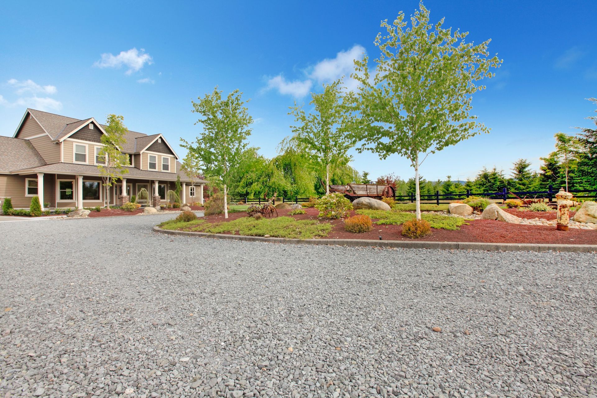 Large gravel driveway beside a suburban house and landscaped yard under a bright blue sky