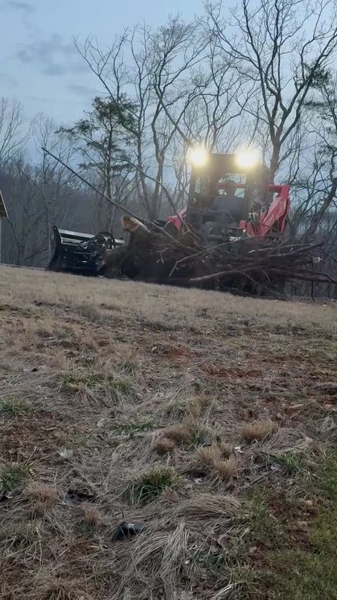 Bulldozer with bright headlights clearing brush in a field at dusk