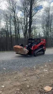 Red skid steer with raised bucket holding a large rock on a gravel road in a wooded area.