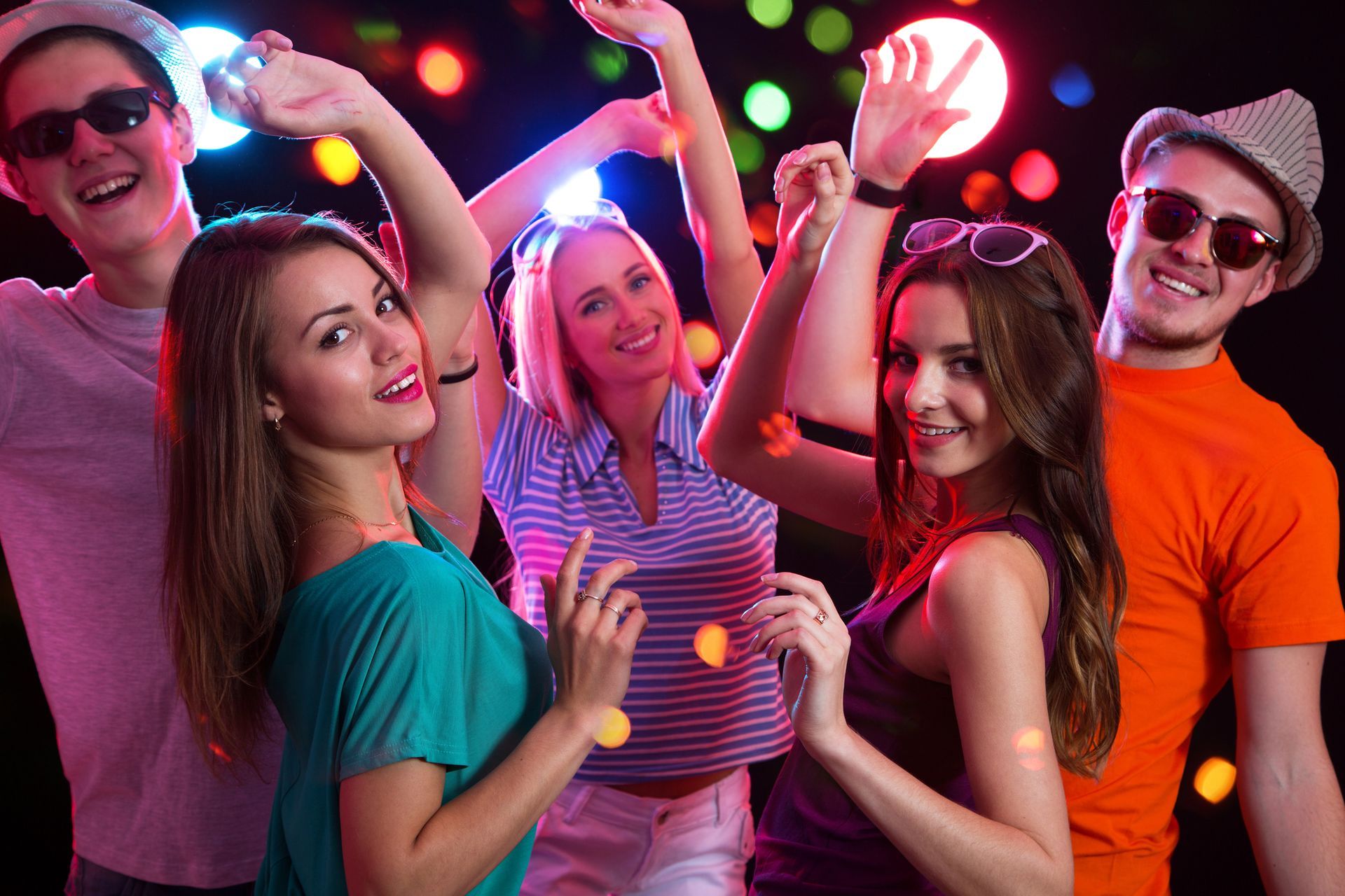 Cinque persone che ballano a una festa con luci colorate; sorridono e tengono le braccia alzate.