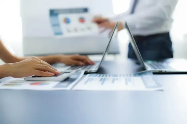 Mani che usano un computer portatile durante una riunione in un ufficio luminoso, con grafici sullo schermo sullo sfondo.