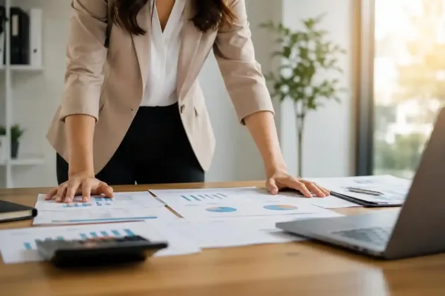 Una persona esamina dei grafici stampati seduta a una scrivania con un computer portatile e dei documenti in un ufficio luminoso.