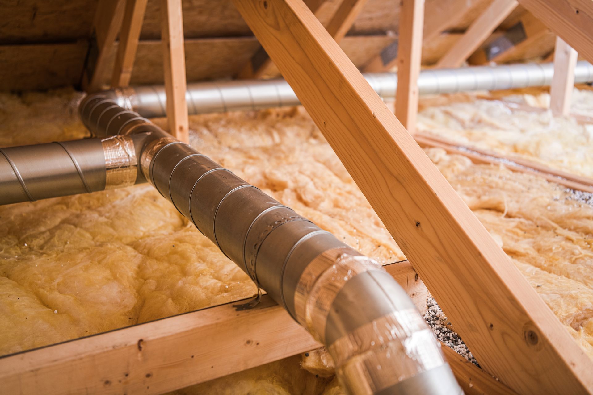 Metal HVAC ductwork in an attic with exposed wooden beams and insulation.