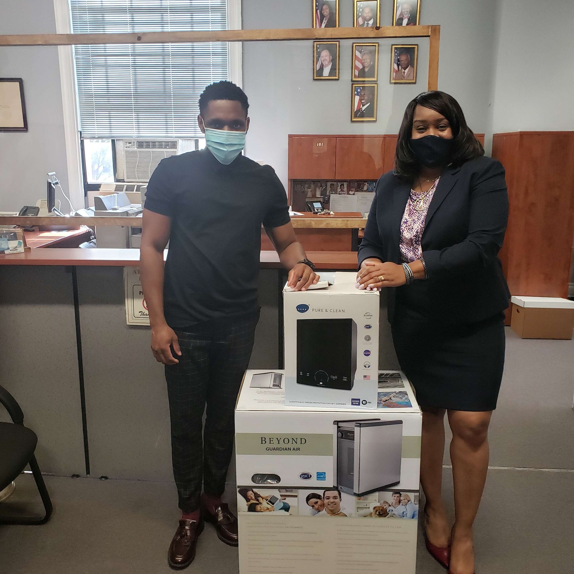 Two masked people stand beside stacked appliance boxes in an office room.