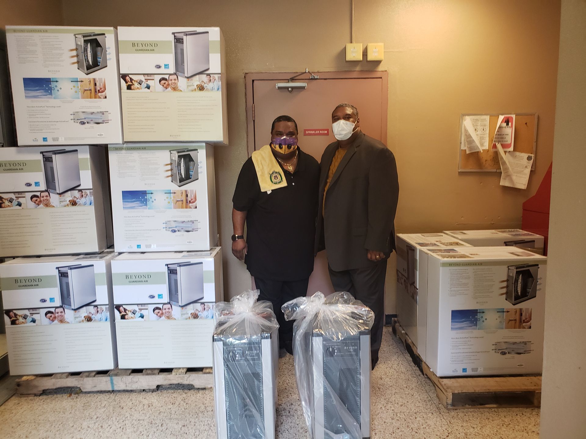 Two masked people stand in a room surrounded by stacked boxes and wrapped supplies.