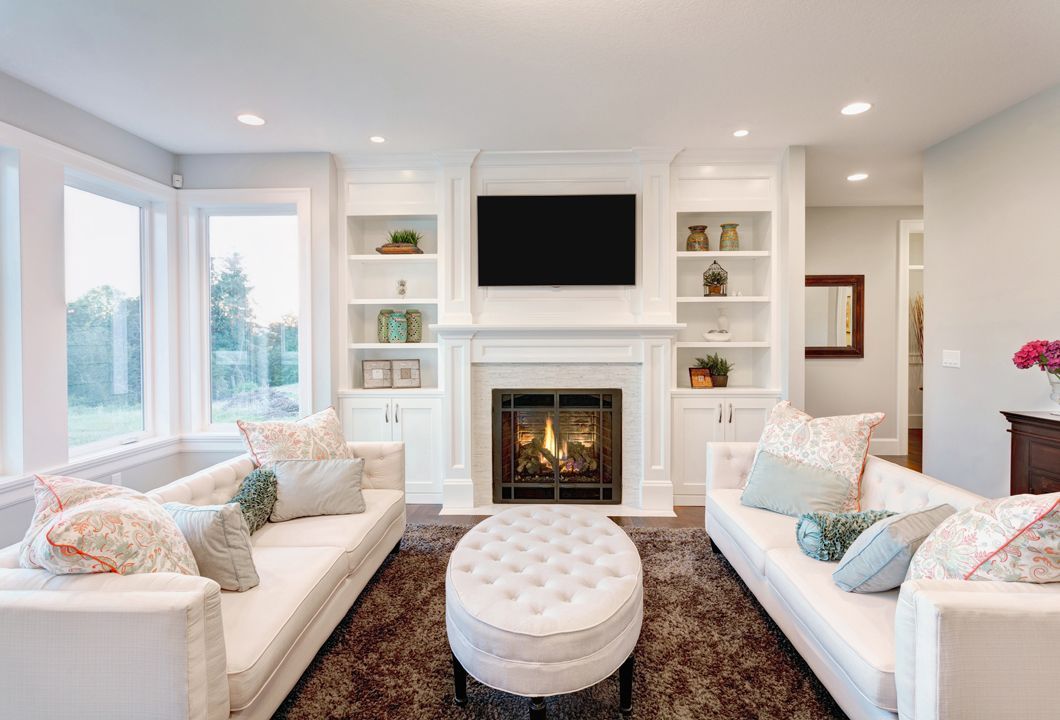 Bright modern living room with white sofas, tufted ottoman, fireplace, built-ins, and large windows