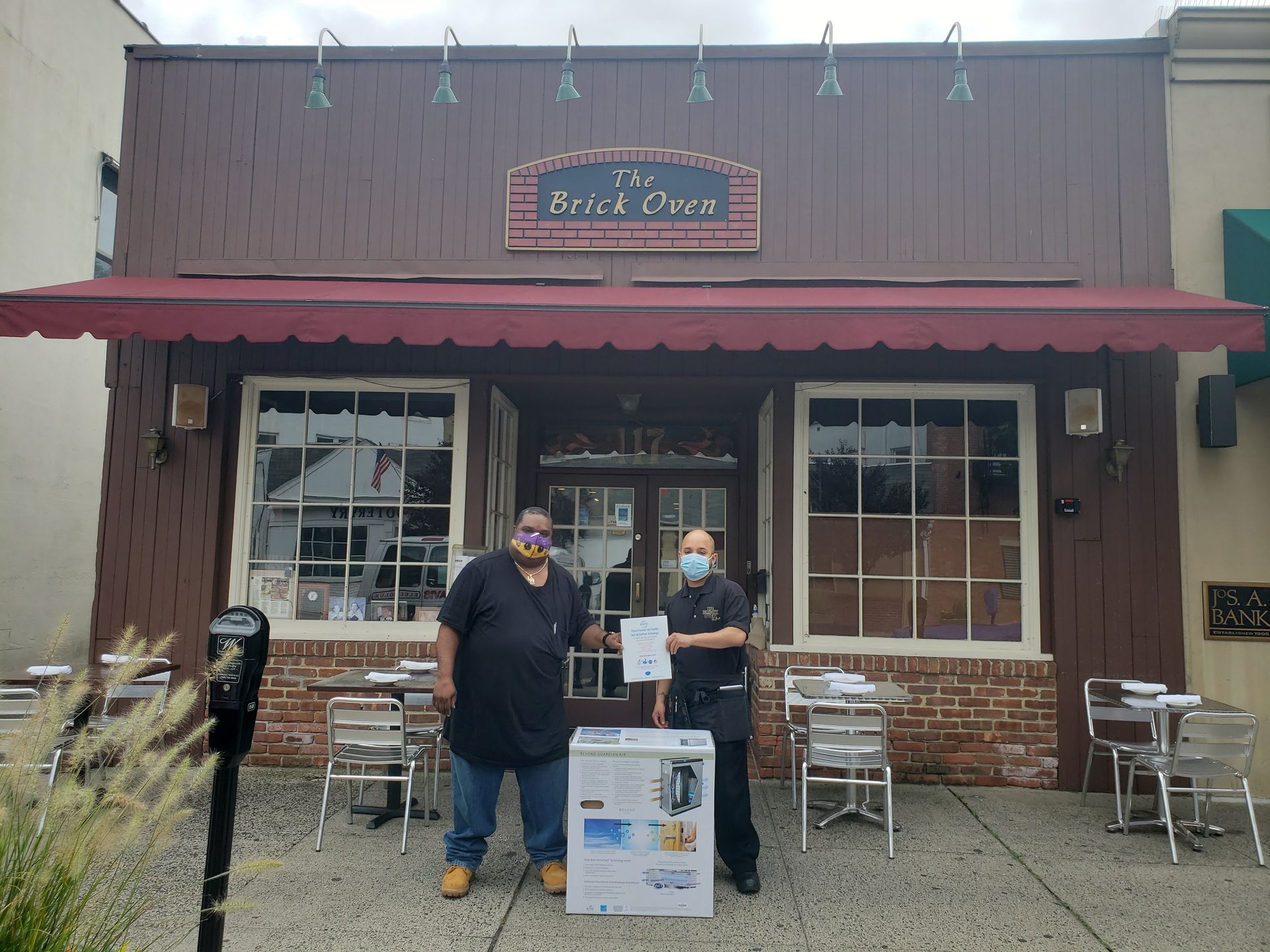 Two people outside a small restaurant with red awning and patio tables, standing by a signboard.