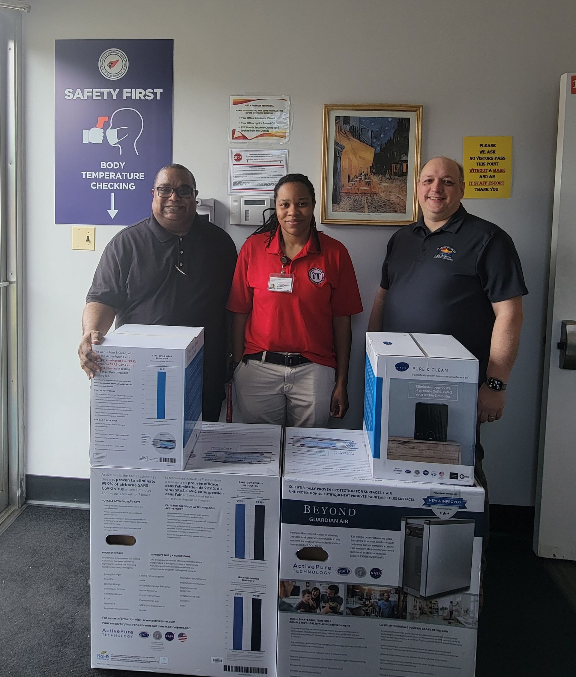 Three people stand behind boxed appliances in a lobby with posters on the wall.