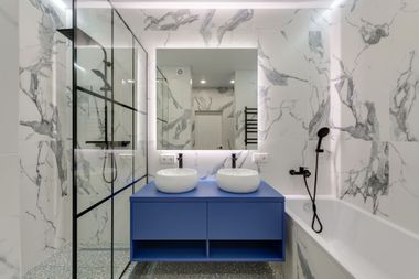 Modern bathroom with marble walls, blue double vanity, mirrors, glass shower, and a bathtub with black fixtures