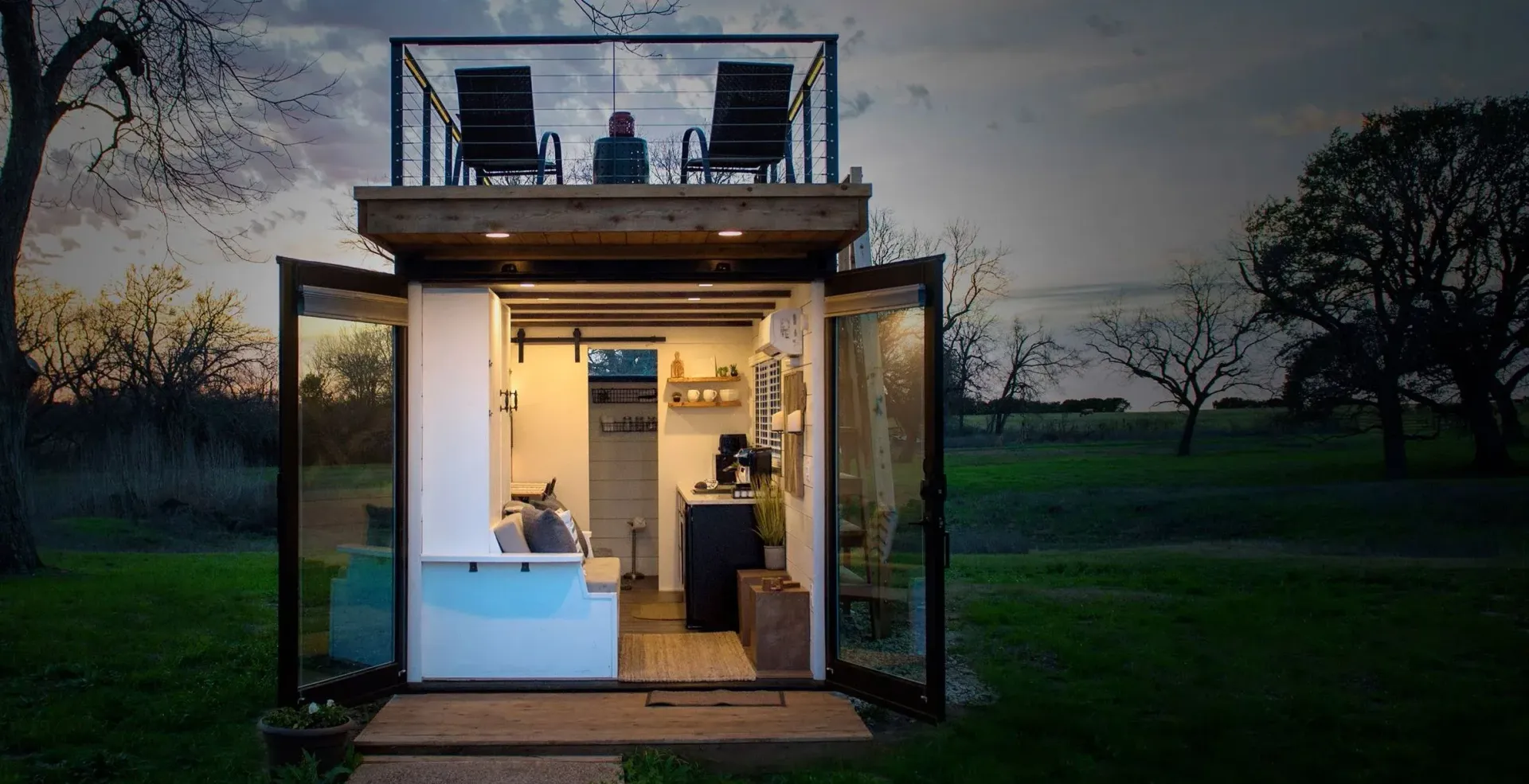 Tiny house with glass doors, open to a green field, a rooftop balcony with two chairs.
