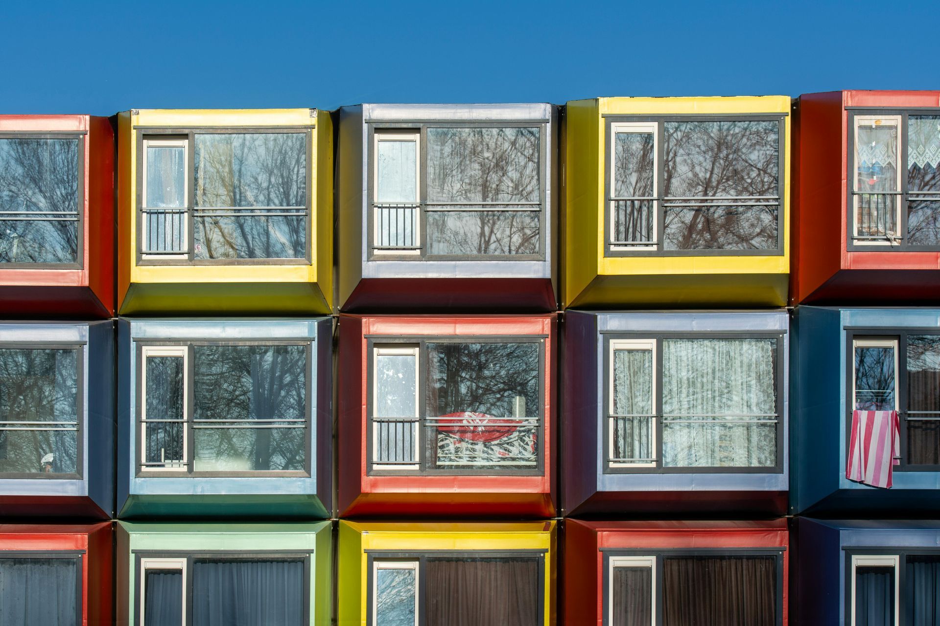 Colorful, stacked modular housing units with windows reflecting trees and sky.