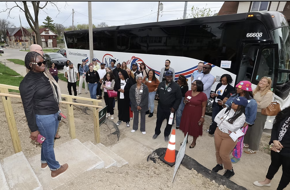 People gathered outside near a black bus, with one person speaking from steps beside a railing.