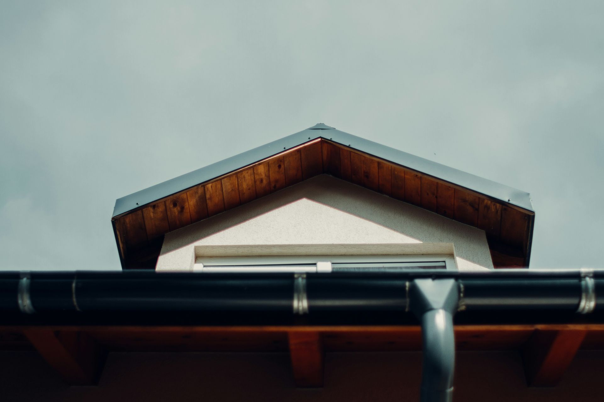 Gabled roof with dark rain gutter and a window against a cloudy sky.