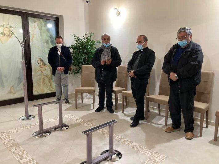 Quattro uomini mascherati sono in piedi in una piccola sala espositiva accanto a opere d'arte incorniciate e supporti per l'esposizione.