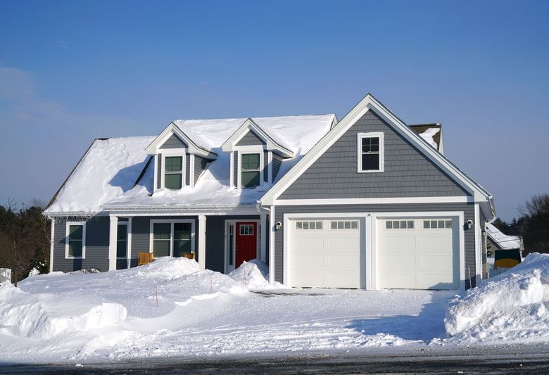Gray house covered in snow with a red front door and white garage doors under a clear blue sky.