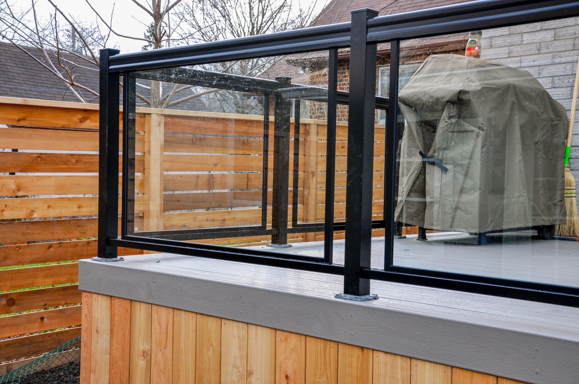 Black framed glass railing on a deck with a covered grill in the background. Wooden fence and siding visible.