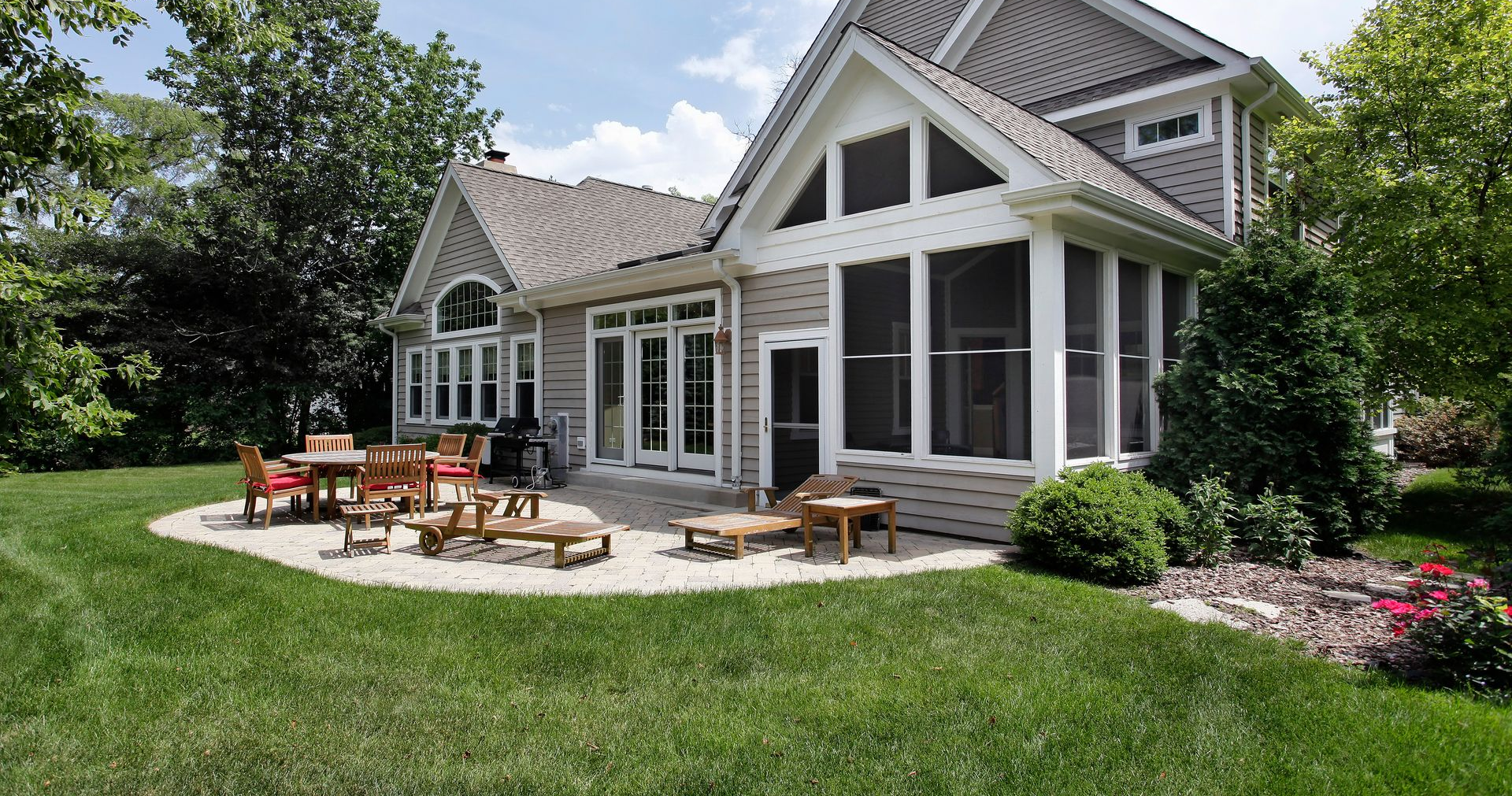 Backyard patio with wooden furniture in front of a house, surrounded by grass and trees.