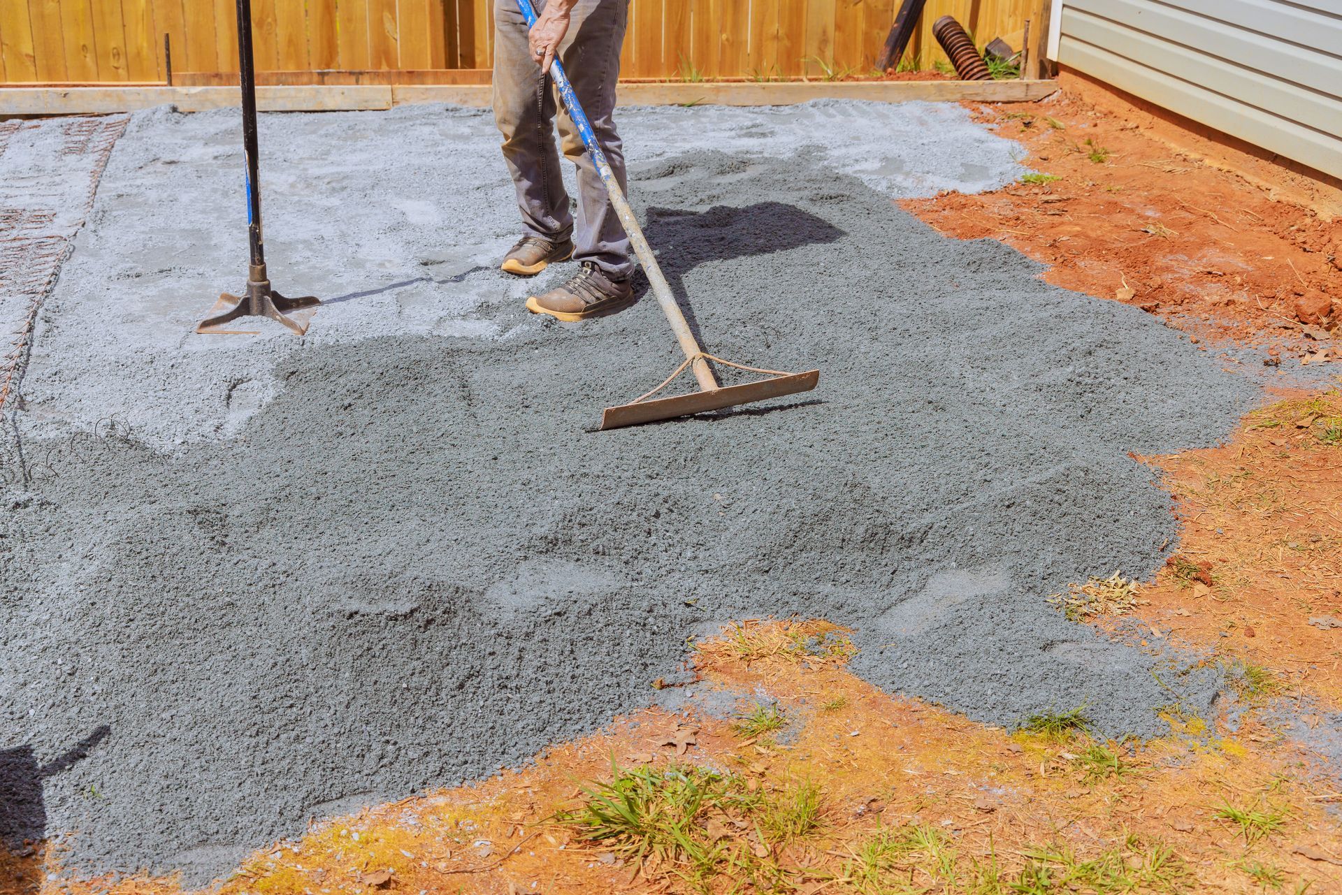 Person rakes crushed stone base for patio, next to a wooden fence on an outdoor worksite.
