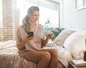A woman is sitting on a bed holding a pill and a cell phone. A woman is sitting on a bed holding a pill and a cell phone.