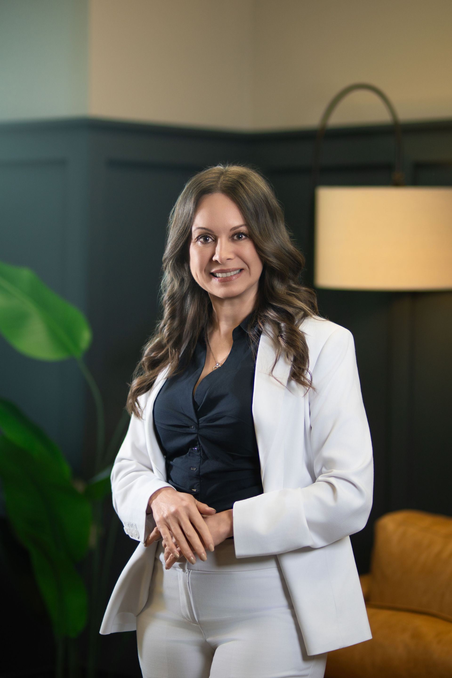 Woman in a white blazer standing indoors, smiling beside a floor lamp and green plant.