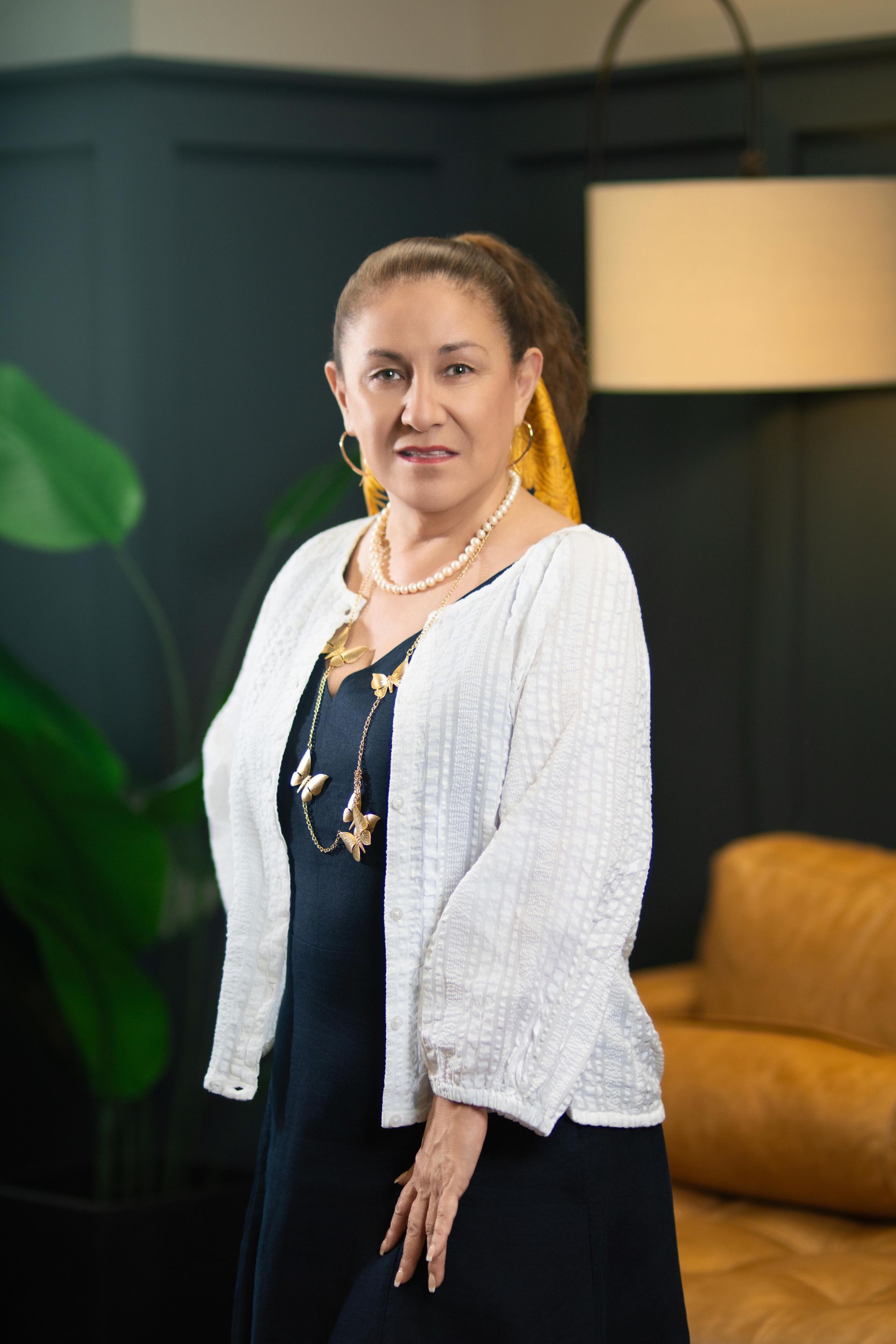Woman standing in a modern room with a lamp and mustard sofa, wearing a white cardigan and navy top.