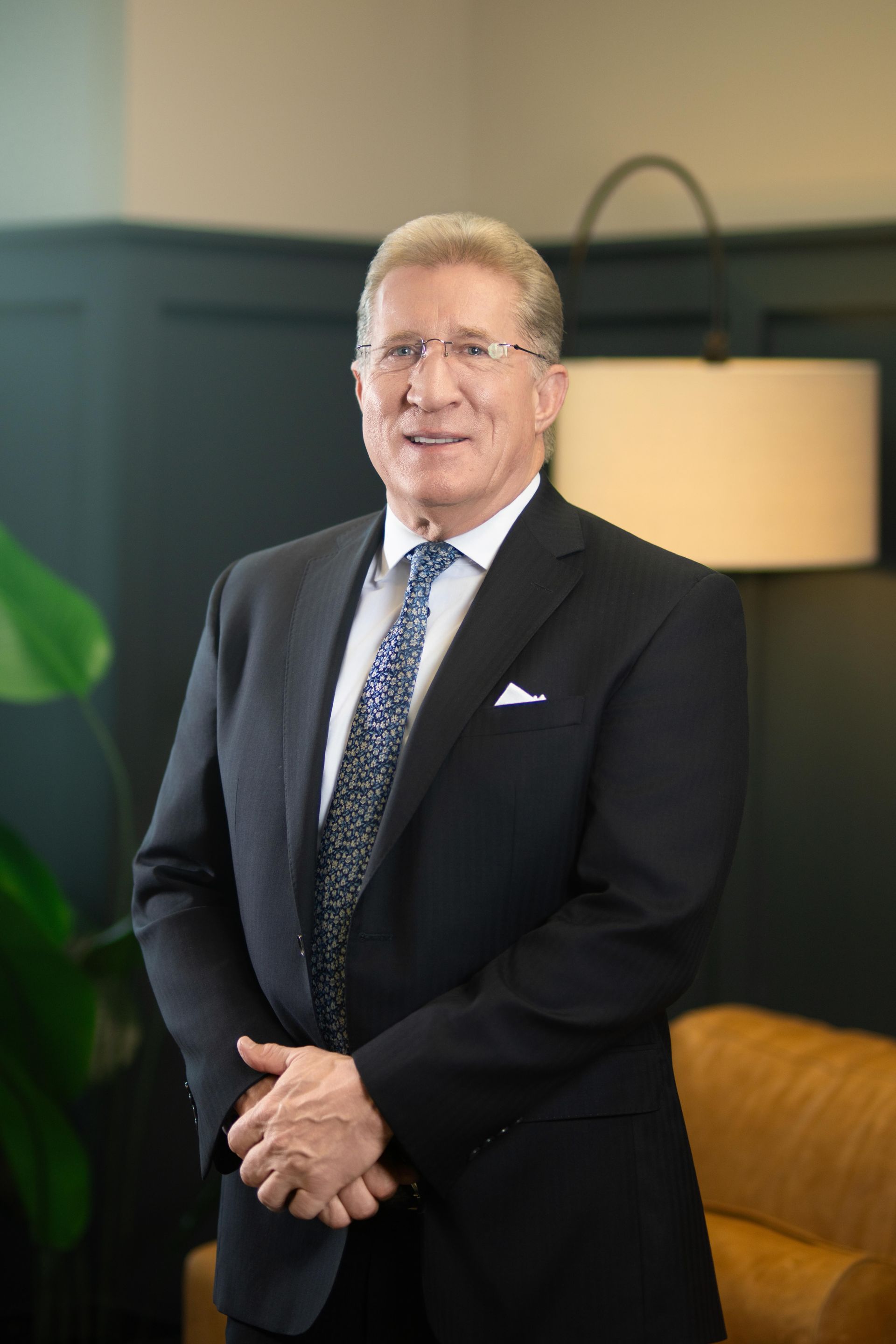 Man in a dark suit standing indoors beside a lamp and tan chair