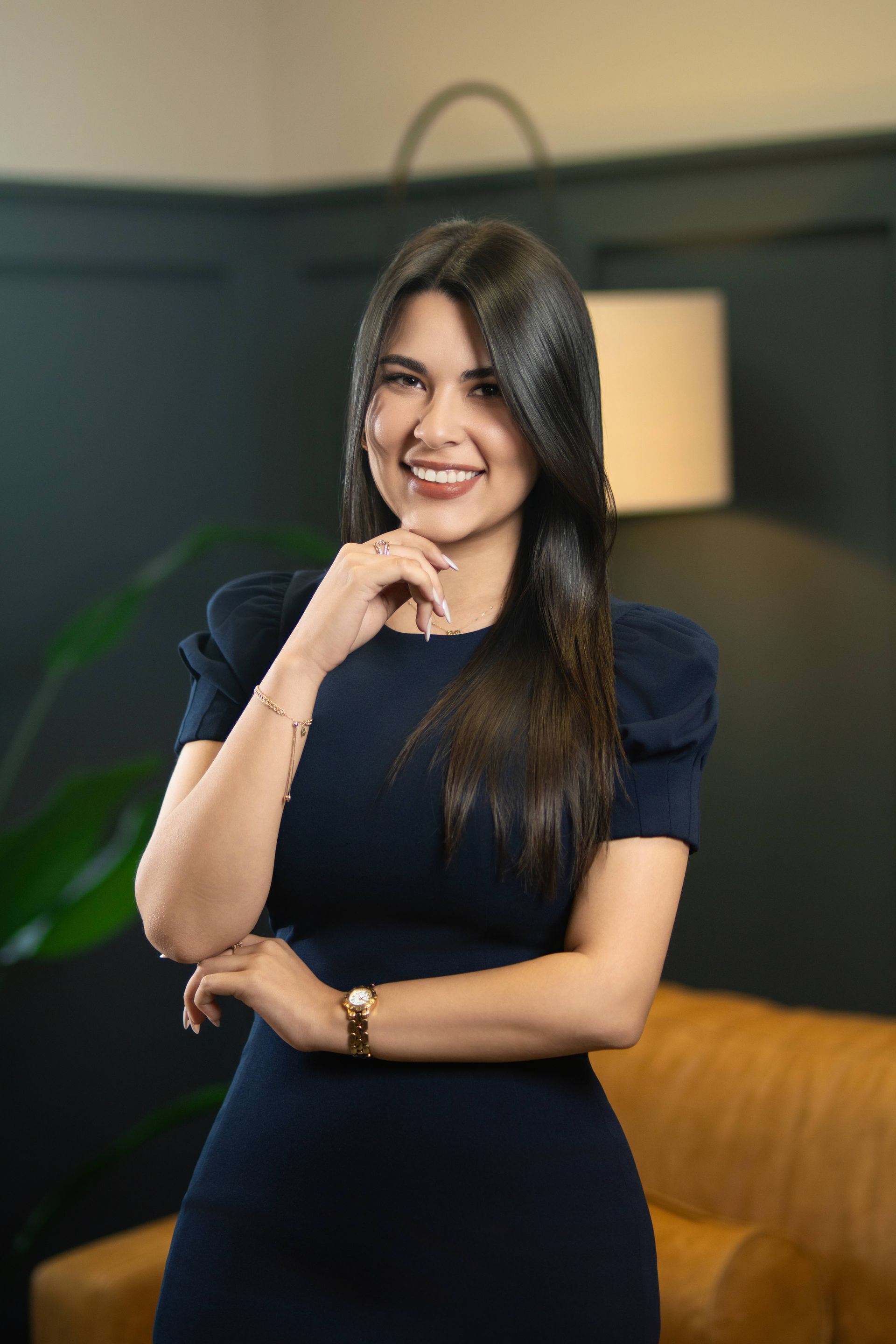Woman in a navy dress smiling with hand on chin in a softly lit room with a lamp and couch.