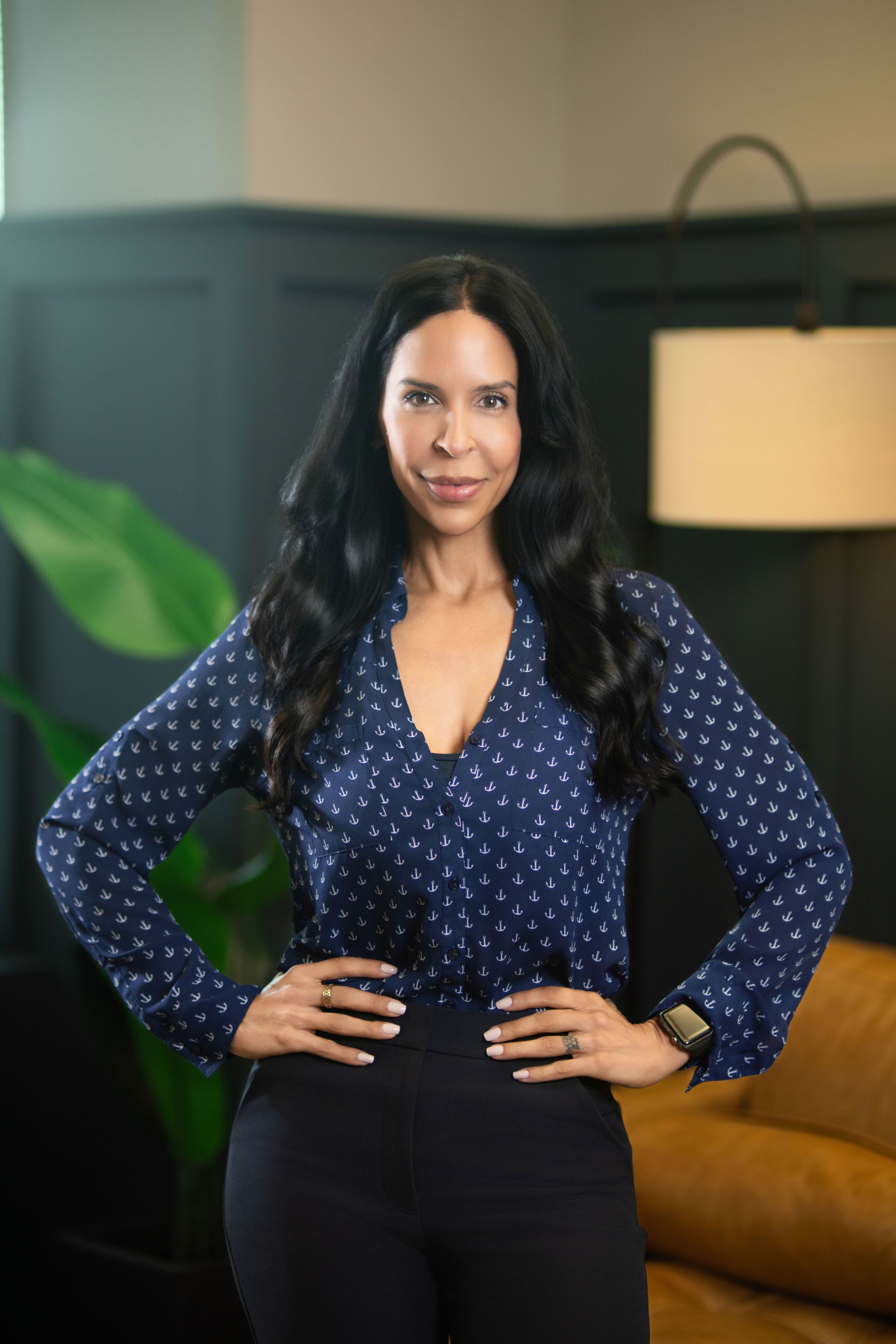 Woman standing with hands on hips in a navy blouse and black pants in a stylish room with a lamp and sofa.