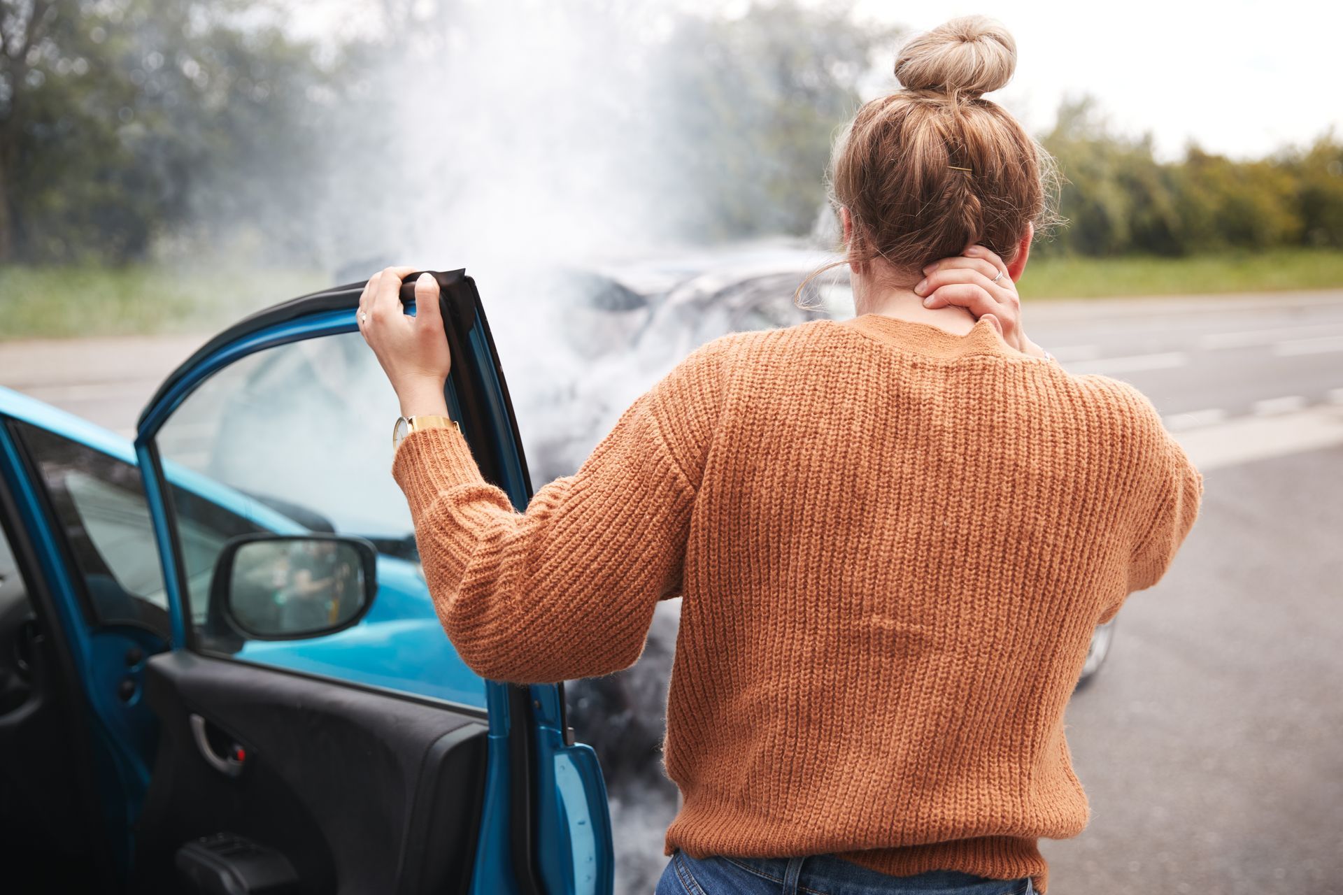 Person in a brown sweater standing by an open car door with steam or smoke billowing from the vehicle in a roadside setting