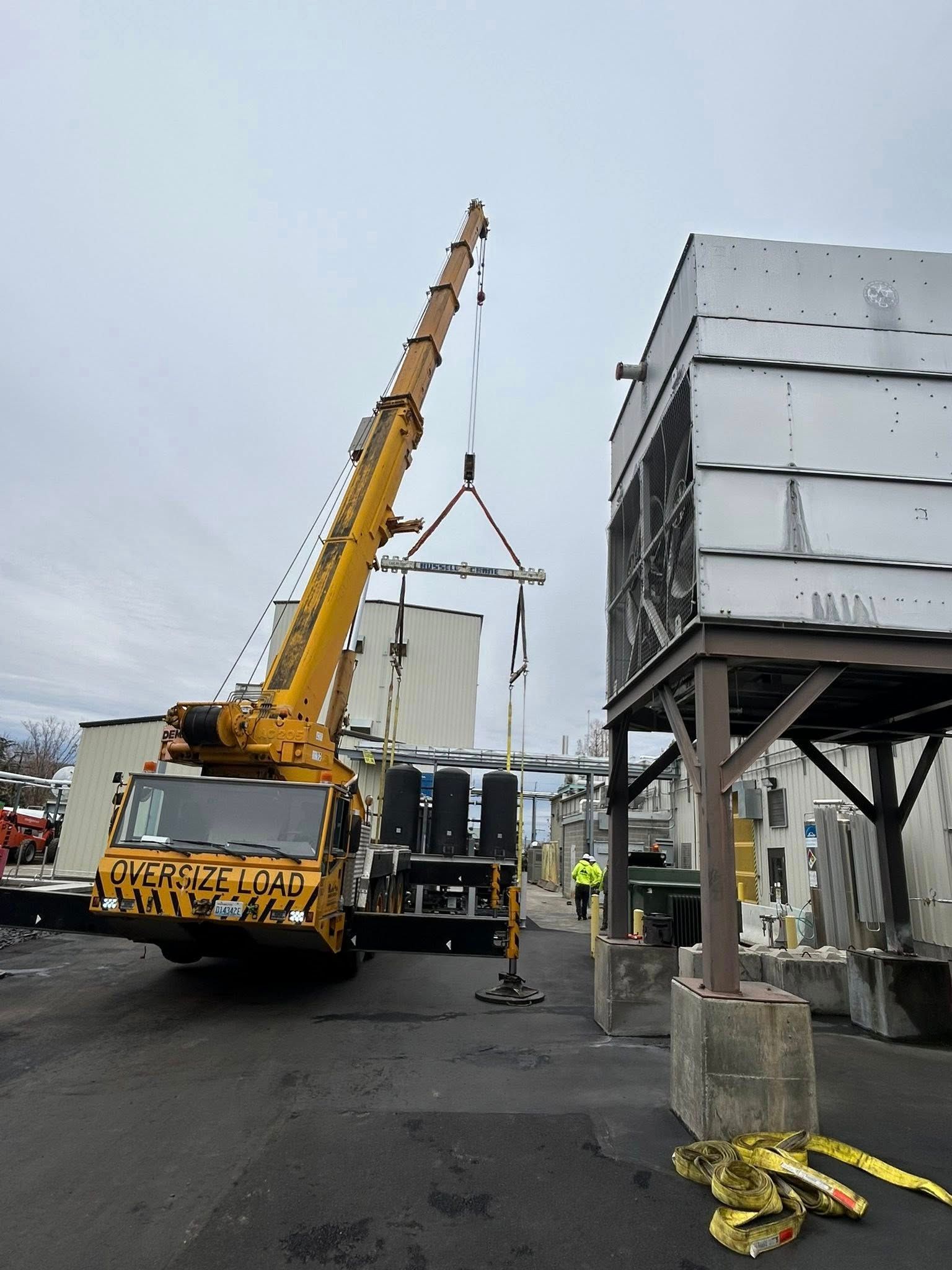 Yellow crane lifting an industrial component next to a large metal structure in an outdoor setting.