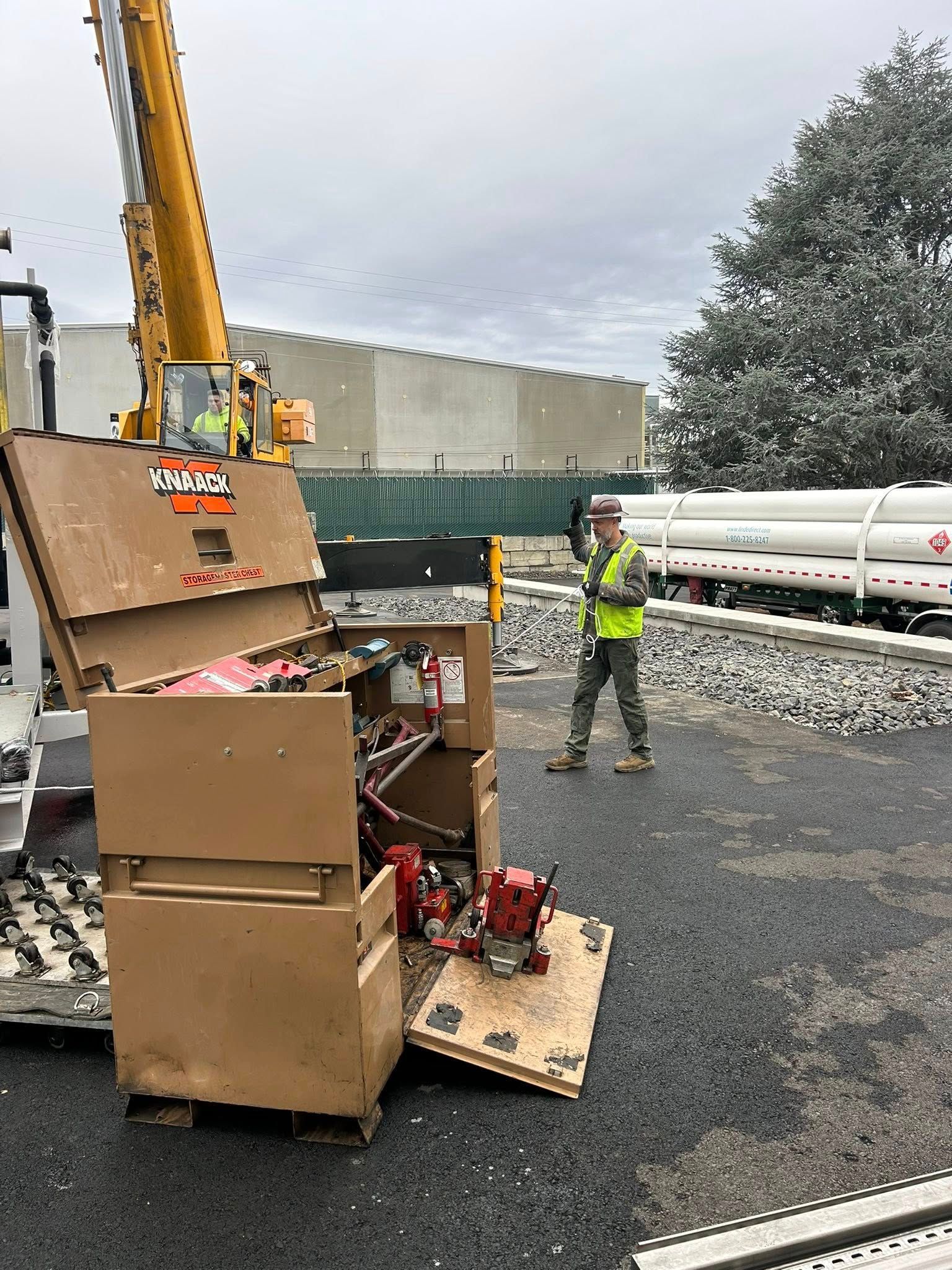 A worker in a vest watches a crane lifting a brown box, near fuel tanks, on gravel.