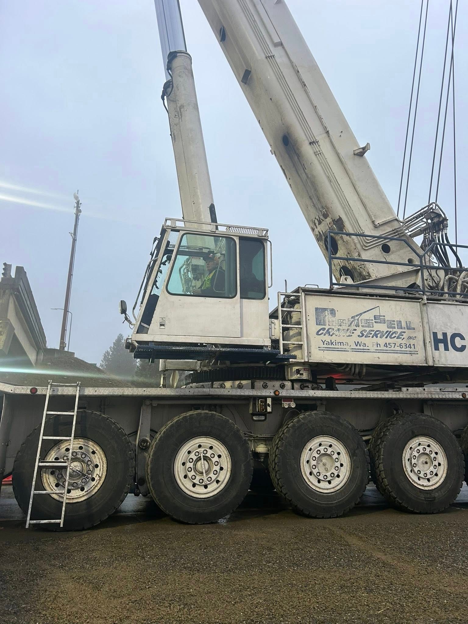 White construction crane on wheels against a gray, overcast sky. Ladder visible.