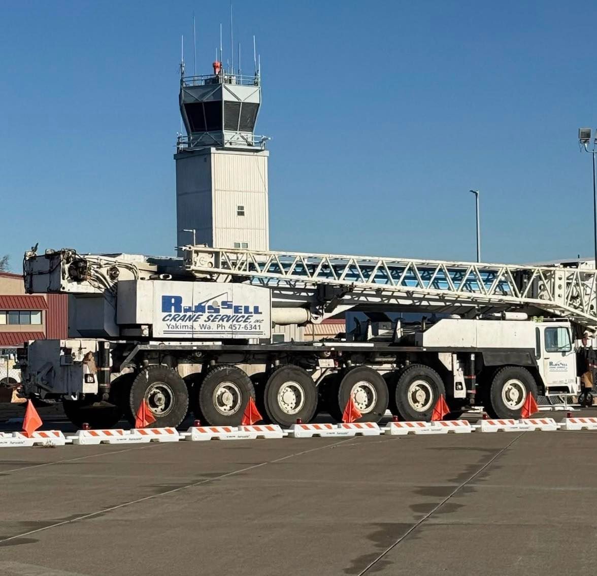 Large white crane parked on a tarmac in front of an airport control tower on a sunny day.