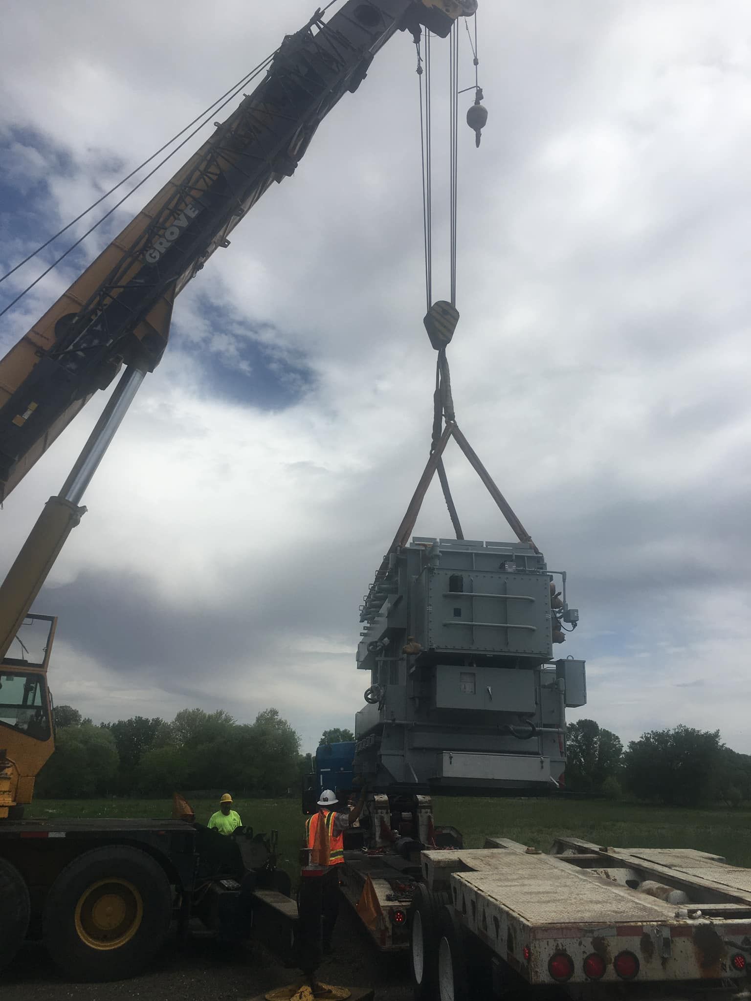 Crane lifting a large electrical transformer from a flatbed trailer, two workers observing.