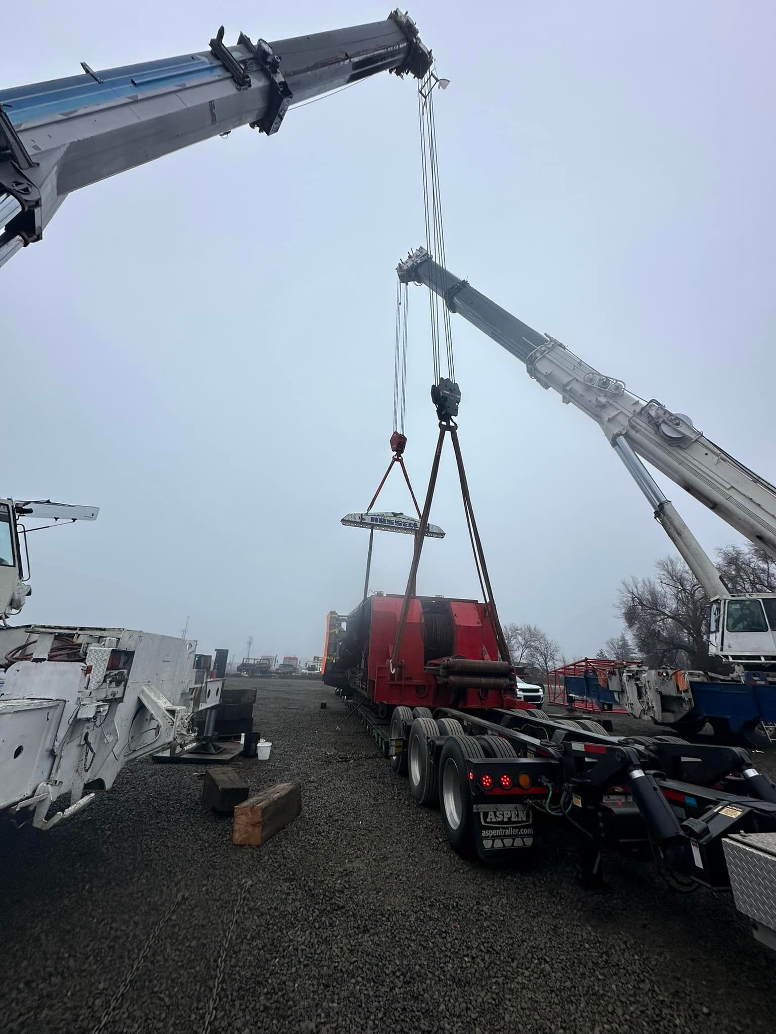 Two cranes lifting a red industrial component onto a truck bed in a foggy outdoor setting.