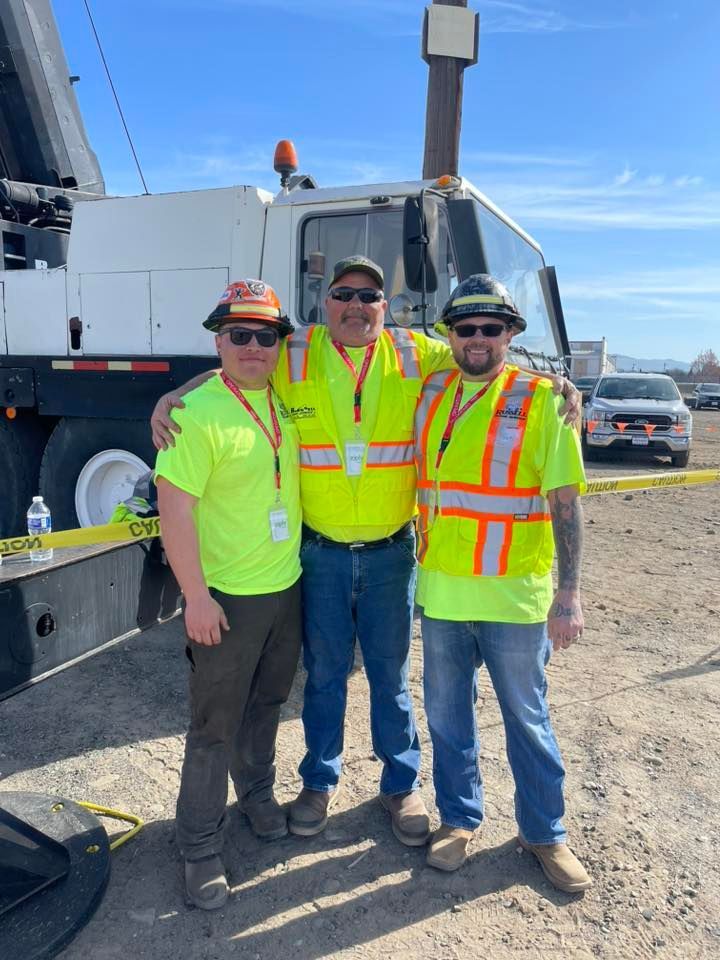 Three construction workers in safety vests and hats pose by a large vehicle on a sunny day.